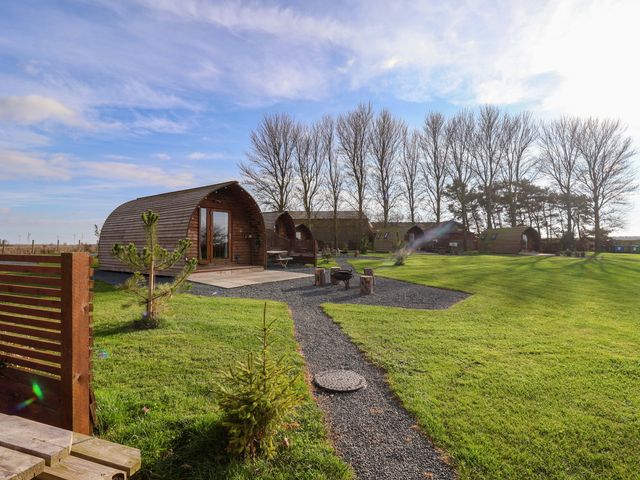 Small wooden cabins on a grassy area with a gravel path and fire pit at Bevy in Little Kelk near Bridlington