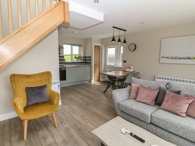 A living room with a yellow armchair a gray sofa with pillows a wooden coffee table and an open kitchen with dining table and chairs at Bwthyn Cae'r Efail in Rhosgoch near Cemaes Bay
