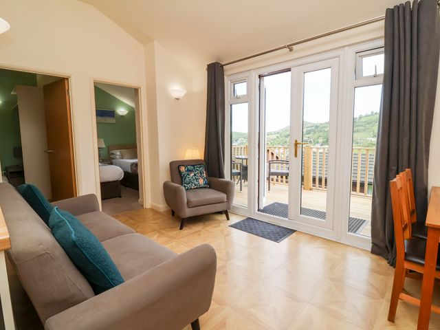 A living room with a sofa, armchair, dining table, and large glass doors leading to a balcony at Chalet Log Cabin L10 in Combe Martin