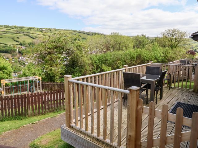 A wooden deck with a table and four chairs overlooking a fenced playground and green hills at Chalet Log Cabin L14 in Combe Martin