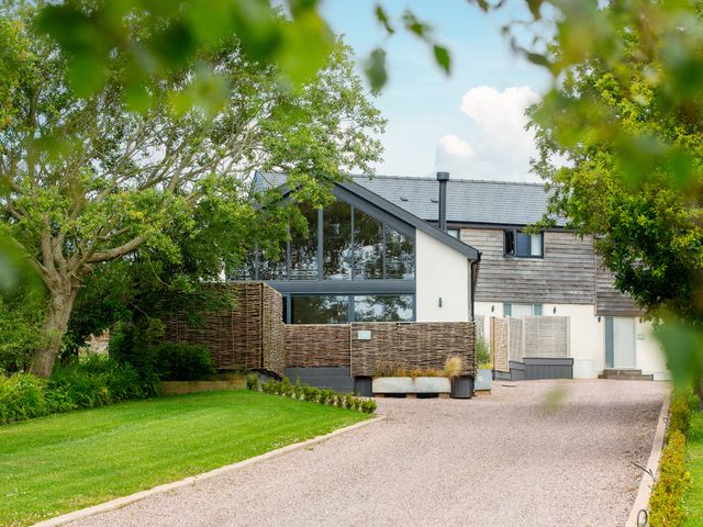A driveway leading to a house with wooden paneling and large windows surrounded by green trees and grass at The Barn in Llanfechell