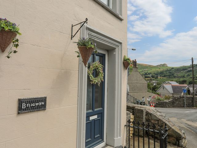 A front door with hanging baskets and a wreath on a wall with a Brigwyn sign and a rural street view at Brigwyn in Nefyn