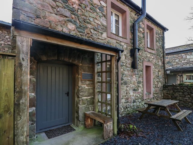 An outdoor stone wall with a grey door under a wooden porch next to a wooden bench and picnic table at Church How Cottage in Nether Wasdale