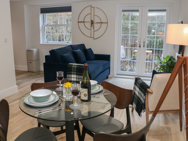 A dining area with a glass table set for two with wine glasses and a bottle and a living room with a blue sofa and a beige armchair next to glass doors at 48 Rock Mill in Stoney Middleton near Calver