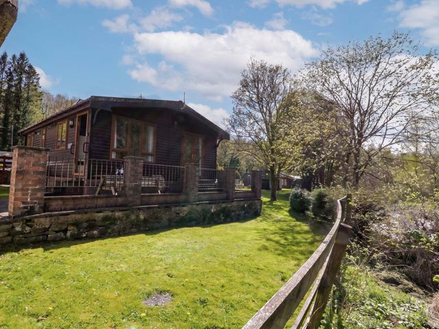 A lodge in a grassy area with trees and a fence at Elm Lodge in Keswick