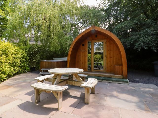 A wooden pod structure with glass door next to a circular picnic table on a stone patio surrounded by trees at Homewood Pod Forton near Garstang