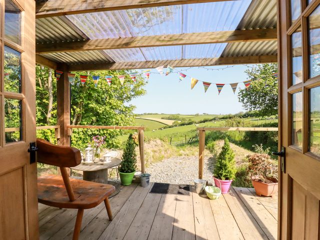 A wooden porch with chair and table surrounded by potted plants and bunting overlooking a rural landscape at Shepherds Retreat in Maxworthy near Crackington Haven