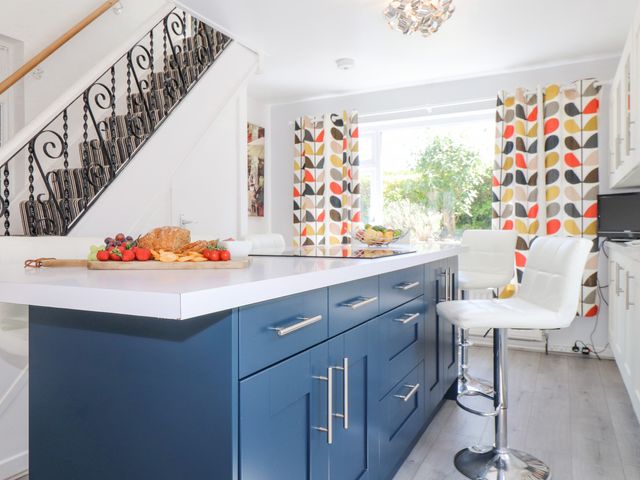 A kitchen with blue island and white chairs near patterned curtains and stairs at Tan Parc in Morfa Nefyn