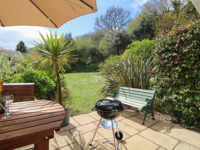A patio with a round wooden table under a large umbrella a charcoal grill a green metal bench and a garden with various plants and trees at Foxglove Cottage in Falmouth