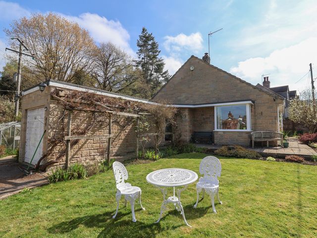 A garden with white metal table and two chairs in front of a stone house with attached garage and trees in the background at Woodstock in Chaffcombe near Chard