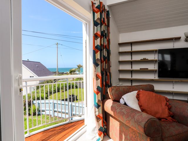 A room with a patterned armchair and cushions next to a glass door leading to a balcony overlooking a garden and the sea at Seaside Sanctuary in Trearddur Bay