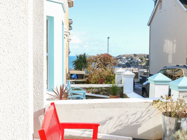 A red bench on a porch overlooking a street with houses plants a parked car and a view of hills and water at The Nest in Brixham