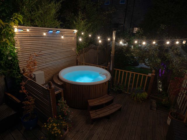 An outdoor wooden deck with a round hot tub illuminated in blue string lights and plants at Rivendell in Ffynnongroyw