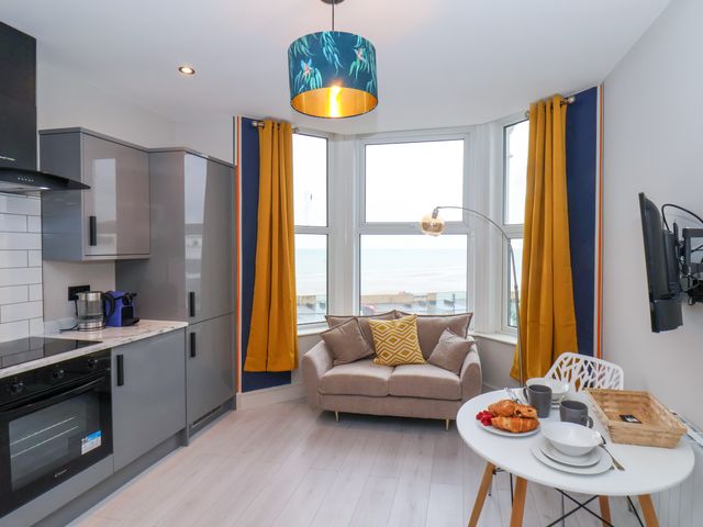 A kitchen and dining area with gray cabinets, a small sofa near windows with yellow curtains, and a table set with croissants and dishes at Apt 6 @ Hunter's Quay in Bridlington