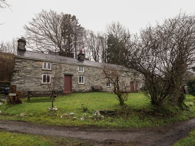 A stone cottage with red doors and white windows in a grassy yard with trees and benches at The Long House in Penmachno