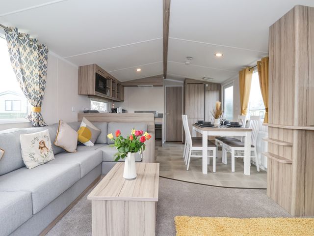 An open plan living room and dining area with a grey sofa and pillows a wooden coffee table with flowers and a dining table with chairs at Lazy Dayz Lodge East Heslerton near Malton