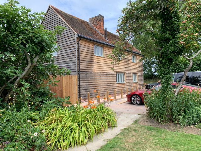 A wooden house with a tiled roof next to a garden with trees flowers and a red car in a driveway at Lodge Cottage in Harwich