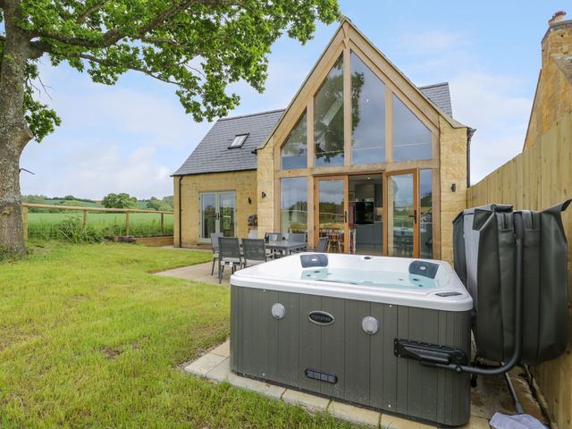 A garden with a hot tub and outdoor dining area next to a stone house with large windows at 4 Wellacres Cottage in Draycott near Moreton-In-Marsh