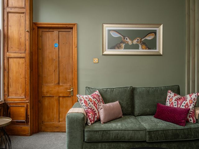 A green sofa with patterned and plain cushions next to a wooden door with a painting of two rabbits on the wall above