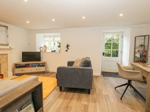 A living room with a television on a stand a gray sofa a desk with a chair and a glass door at Half Pint Cottage in Beaminster