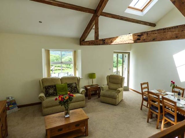 A living room with two armchairs a coffee table with flowers a dining table and wooden beams at Ghyll Bank Cow Shed in Staveley near Windermere