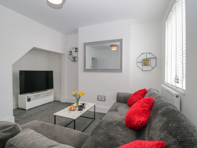 A living room with a gray sofa with red cushions a white coffee table with flowers and croissants a flat-screen TV on a white stand and wall shelves with plants at Glenmore in Plymouth