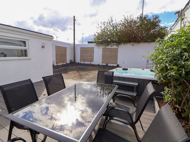 An outdoor patio with a glass table and six black chairs a hot tub and white walls with lattice panels at The Railway Cottage in Llandudno