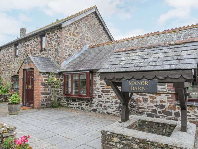 Stone house with a wooden door and windows next to a stone well with a roof and manor barn sign at Manor Barn Saltash