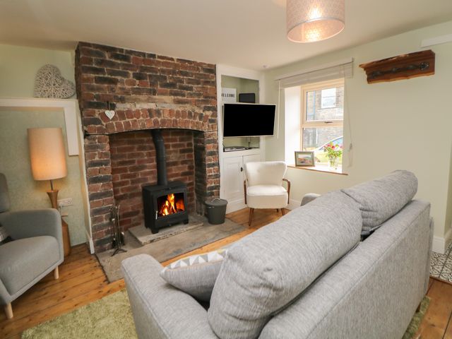 A living room with a brick fireplace wood stove a gray sofa a white armchair a wall-mounted television and a window at The Cottage Bradwell in Bradwell