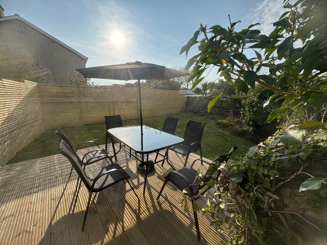 An outdoor deck with a glass table and six chairs under an umbrella with a wooden fence and garden at 1 Mona Terrace in Llanfachraeth