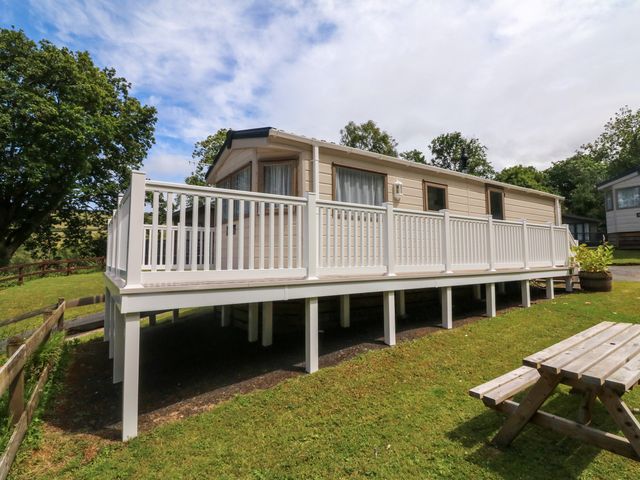 A mobile home with white railings on a raised platform surrounded by grass and trees at 23 Manleigh Park in Combe Martin