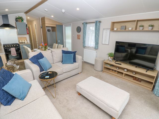 A living room with sofas and blue cushions a television on a wooden stand and a dining area and kitchen in the background at Serenity Thorness Bay near Cowes