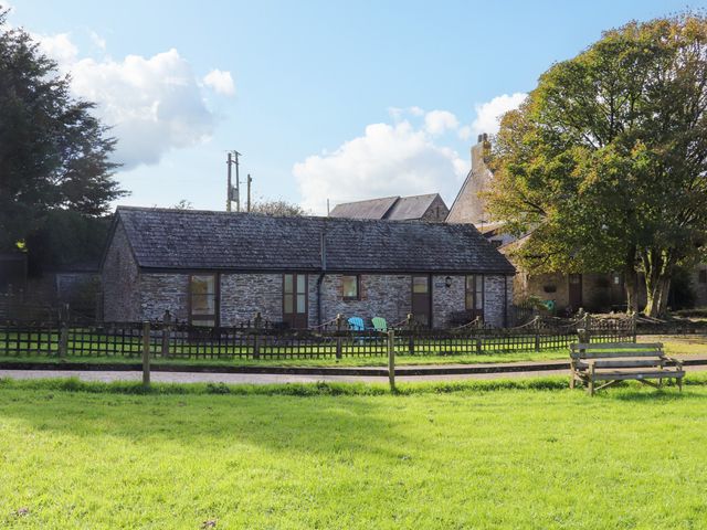A stone building with a pitched roof behind a wooden fence and green grass with a wooden bench at The Mealhouse in Pelynt near Looe