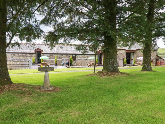 A green lawn with a birdbath and trees in front of stone buildings with picnic tables and umbrellas at The Byre in Common Moor near Liskeard