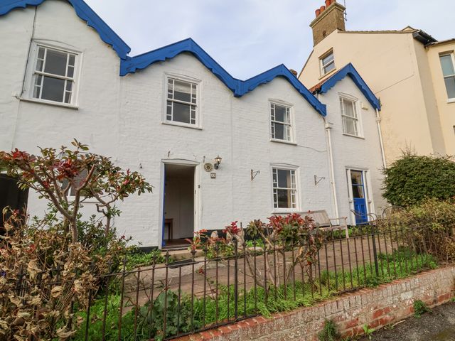Exterior view of white brick houses with blue trim and a small front garden with plants and a metal fence at Iona Cottage in Southwold
