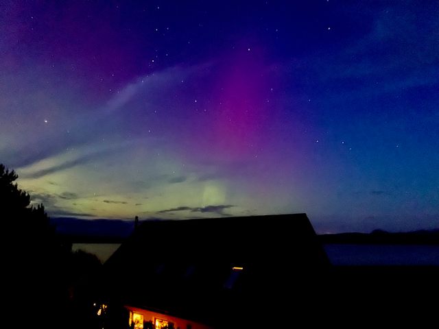 A night sky with stars and northern lights above a dark building near a body of water at Calmdale in Inverasdale near Poolewe
