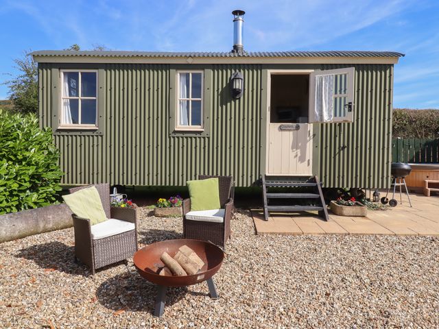 A green metal shepherd hut with two windows and an open door outdoor seating with cushions a fire pit and a barbecue at Shepherds Joy Kennerleigh near Cheriton Fitzpaine