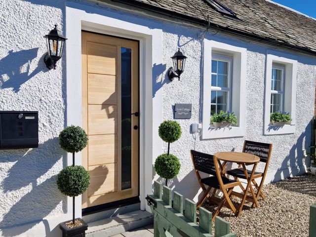 The entrance of a white cottage with a wooden door two black wall lanterns a small round wooden table with two chairs and two topiary plants at Lyne Cottage The Retreat in Romanno Bridge