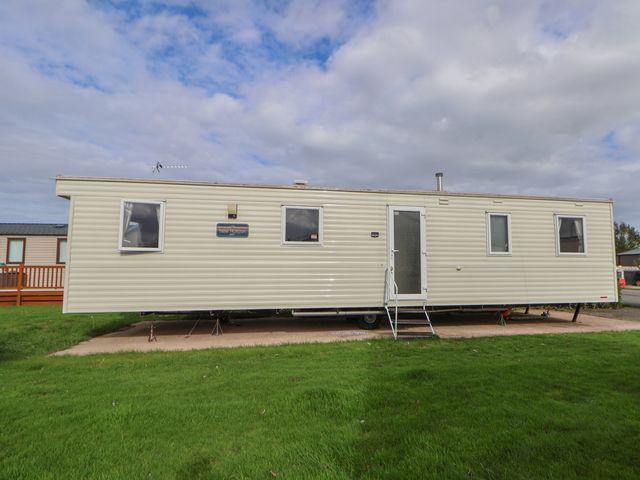 A beige mobile home with four windows and a door with steps on a grassy plot at Herrylock in Rhyl