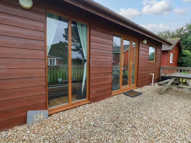 A wooden cabin exterior with glass doors and windows and a picnic table on gravel at Gwanda Tregolds near St Merryn