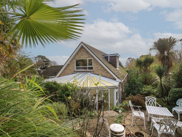 A garden with white patio furniture surrounded by plants and a house with a conservatory at Tall Timbers in Ventnor