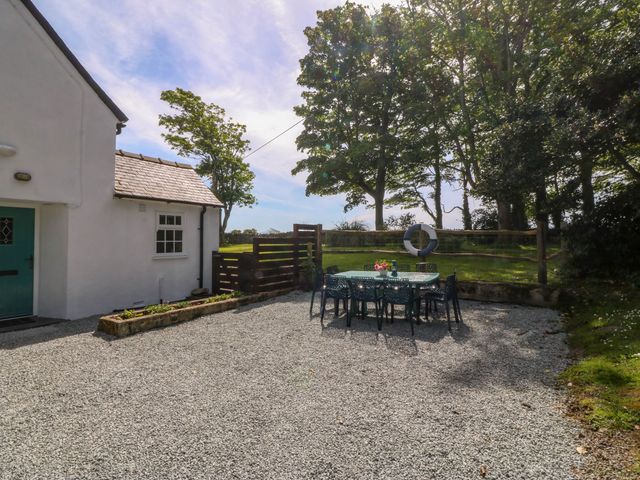 An outdoor patio area with a table and chairs next to a white house and trees at Bodior Garden Cottage in Rhoscolyn