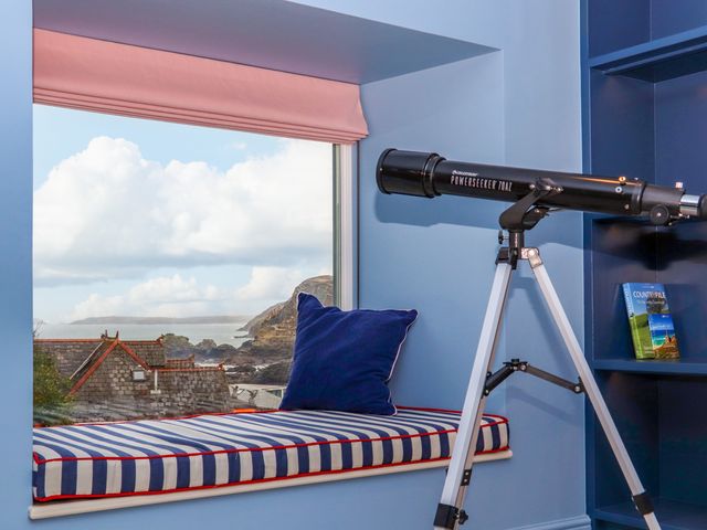 A telescope on a tripod next to a window seat with a striped cushion and blue pillow at Mavericks Wave in St Agnes