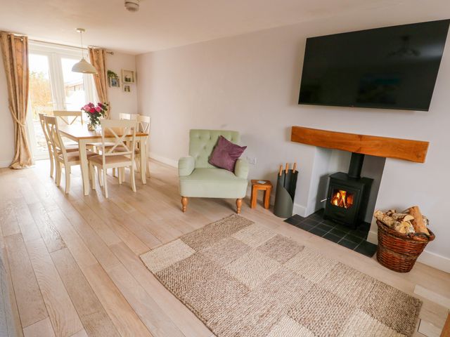 A dining area with a wooden table and chairs next to glass doors a green armchair with a purple pillow a wood burning stove with a wooden mantel and a basket of logs a flat screen tv mounted on the wall at Ty Bach in Llanfaelog near Rhosneigr