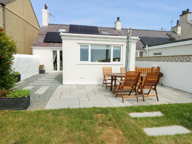 A backyard patio with wooden table and chairs on stone tiles beside a white house at Ty Bach Llanfaelog near Rhosneigr
