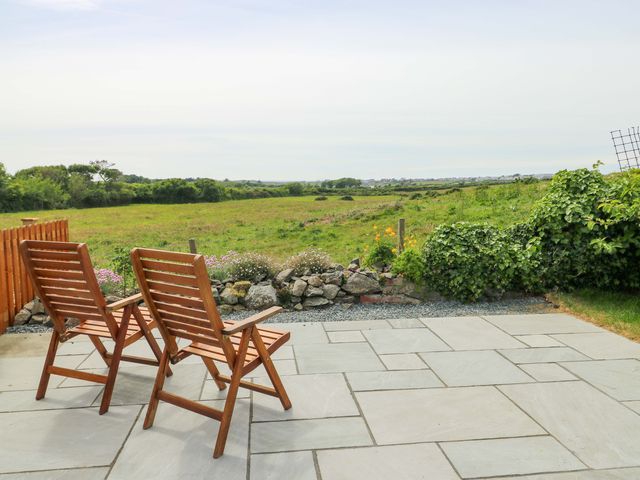 Two wooden chairs on a stone patio overlooking a grassy field with flowers and shrubs at Ty Bach in Llanfaelog near Rhosneigr