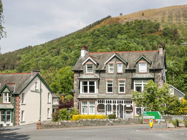 A large stone house with multiple windows and chimneys next to a road with directional signs in front of a forested hill
