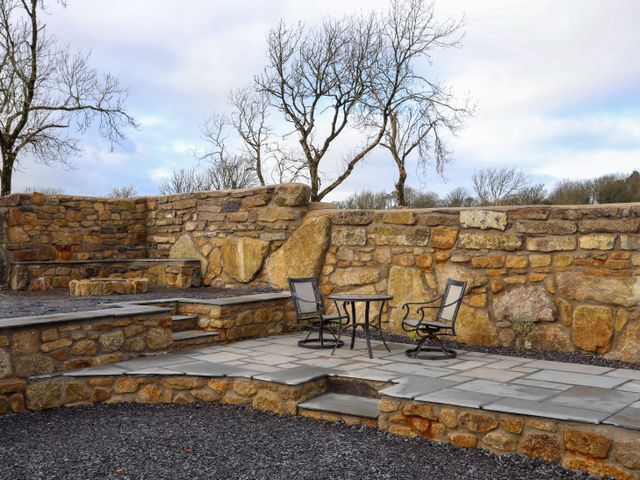 An outdoor stone patio area with a table and two chairs surrounded by a stone wall with trees in the background at Ynys Uchaf Near Benllech