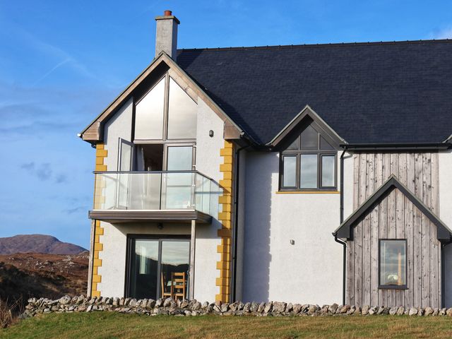 A modern house with large windows and a glass balcony with a stone wall and grassy hill outside at Riverside in Lingerbay near Leverburgh