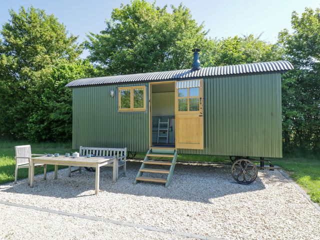 A green corrugated metal shepherd hut with open door and window on a gravel patio with wooden table chairs and bench surrounded by trees at Hazel South Leigh near Witney